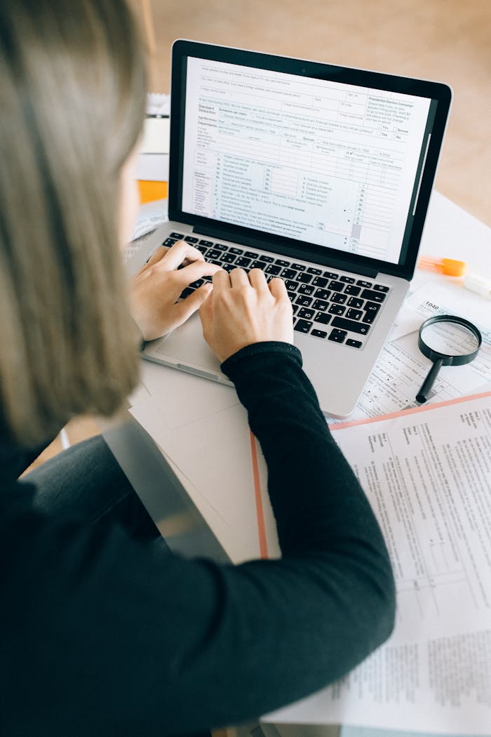services-03 Focused woman working on laptop in office, surrounded by paperwork and magnifying glass.